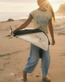 A woman walking barefoot on a sandy beach at sunset, carrying a white surfboard and a black wetsuit or wet top. She is wearing an Atmosea shirt with blue trim and light blue cropped pants.