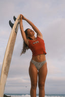 A woman posing next to a surfboard on the beach in the Inner Relm Checkers Tank and bikini bottoms. 