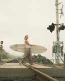 A woman carrying a white surfboard walks across railroad tracks in an overcast outdoor setting, wearing a white tank top and Atmosea pants. 