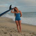 A woman on the beach wearing the Seea Rosita one piece in ultramarine, posing with a surf board