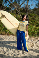 Woman holding a surfboard and wearing the Atmosea apres surf pants in blue and yellow on a sandy beach with trees in the background