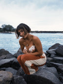 A model at the beach crouching and wearing the Atmosea Après Surf Panel Pants in rust brown cotton terry with cream side panels, a relaxed wide-leg silhouette, and adjustable drawstring waist.