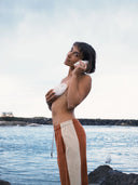 A model at the beach wearing the Atmosea Après Surf Panel Pants in rust brown cotton terry with cream side panels, a relaxed wide-leg silhouette, and adjustable drawstring waist.