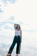 A model posing at the beach wearing the Atmosea Après Surf Pants in forest green cotton terry towelling with a relaxed wide-leg fit, elastic drawstring waist, and sporty canvas side stripes.