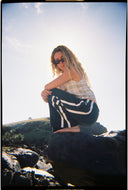 A model on a rock wearing the Atmosea Après Surf Pants in forest green cotton terry towelling with a relaxed wide-leg fit, elastic drawstring waist, and sporty canvas side stripes.