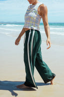A side view of a model at the beach wearing the Atmosea Après Surf Pants in forest green cotton terry towelling with a relaxed wide-leg fit, elastic drawstring waist, and sporty canvas side stripes.