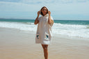 A model outside looking in the distance and wearing the Atmosea Delfi Après Surf Robe made from plush cotton terry towelling with a zip front, oversized hood, front pockets, and dolphin embroidery.