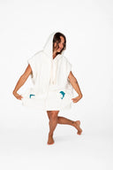 A model in the studio showing the Atmosea Delfi Après Surf Robe made from plush cotton terry towelling with a zip front, oversized hood, front pockets, and dolphin embroidery.