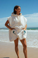 A model at the beach wearing the Atmosea Delfi Après Surf Robe made from plush cotton terry towelling with a zip front, oversized hood, front pockets, and dolphin embroidery.