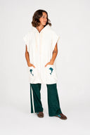 A model wearing the Atmosea Delfi Après Surf Robe made from plush cotton terry towelling with a zip front, oversized hood, front pockets, and dolphin embroidery in a studio. 
