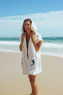 A model outside wearing the Atmosea Delfi Après Surf Robe made from plush cotton terry towelling with a zip front, oversized hood, front pockets, and dolphin embroidery.