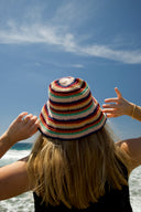A picture of the back of a woman wearing the Atmosea rainbow crochet hat at the beach on a sunny day. 