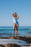 Model wearing the Inner Relm Poolside One Piece standing on coastal rocks by the ocean.