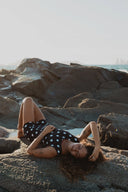 Model relaxing on coastal rocks wearing the Our Ripples Canda Surfsuit 0.8mm.