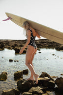 Model carrying a surfboard while wearing the Our Ripples Kiki Suit 0.8mm on a rocky shoreline.