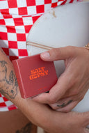 Hand holding the Salt Gypsy Premium Eco Surf Wax - All Water in biodegradable packaging beside a surfboard.