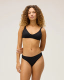 The front view of a model wearing the Seea Phoebe Bikini Bottom in Black featuring textured recycled fabric, high-cut hips, and cheeky coverage.