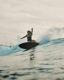 A woman surfing on a short board in the  Seea Rosita One Piece swimsuit in Black featuring white contrast trim, adjustable straps, and a low scoop back.