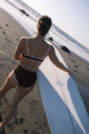 A woman holding a surfboard and wearing the September cold brew top from behind