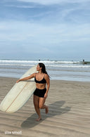 A model outside wearing the September Maya Bikini Top in Midnight Black Terry and carrying a surfboard 