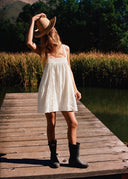 A model ourside on a dock wearing the Sisstr Casey Mini Woven Tank Dress in Sea Salt, featuring a square neckline, embroidered details, and lightweight cotton fabric.