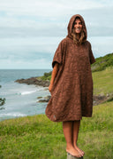 Model wearing the Sunward Bound Hooded Changing Towel in Cacao standing on a grassy coastline overlooking the ocean.