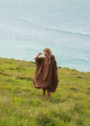 Model wearing the Sunward Bound Hooded Changing Towel in Cacao walking along a grassy bluff above the ocean.