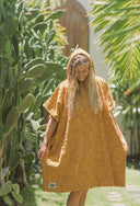 Model wearing the Sunward Bound Hooded Changing Towel in Turmeric standing outdoors among tropical greenery.
